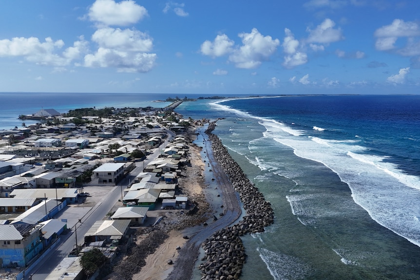 A wall of rocks at the shore of a thin strip of land with buildings on it, surrounded by ocean and a lagoon.