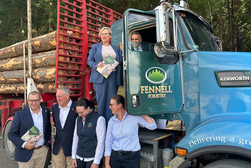 four people standing in front of a truck with logs on the trailer, with one person standing on the truck and another in the cab