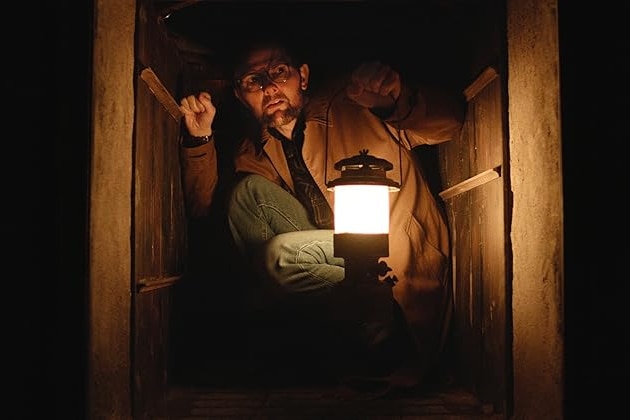 A man crouches in a small, dark space with a lantern