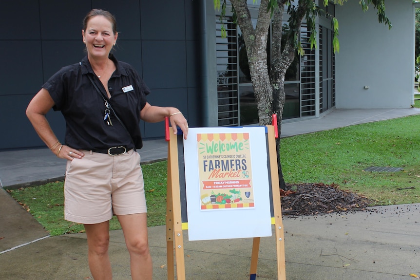 A female teachers stands beside a sign welcoming people to the farmers market