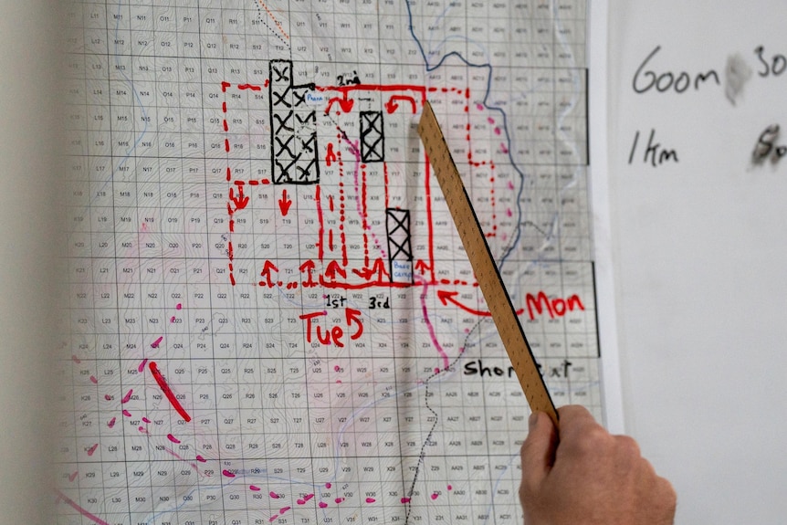 A person holding a wooden ruler points at a white map on the wall. Red and black markings indicate movements and direction