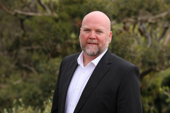 A man wearing a suit smiles in front of a blurred background of trees.