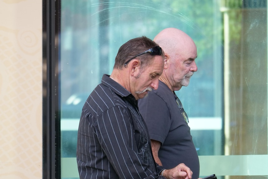 A middle-aged man with a moustache and a bald man walk side by side past a court building.