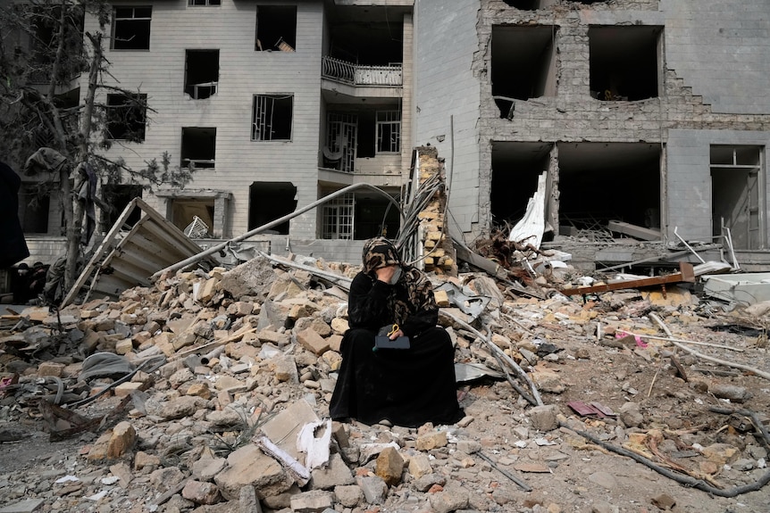 A woman holds her head while sitting in rubble in front of destroyed apartment buildings.