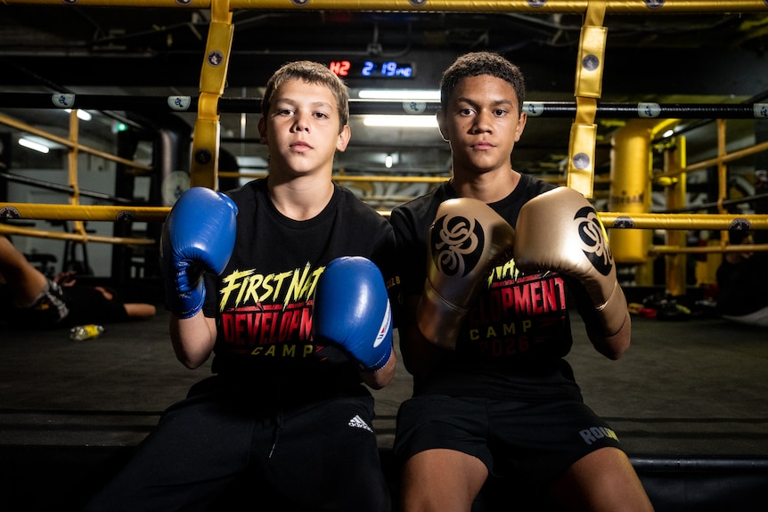 Two young boys sit side-by-side wearing boxing gloves in a gym.