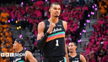 Victor Wembanyama, wearing a black San Antonio vest with a turquoise, pink, and orange stripe across the chest, punches his right fist in celebration and smiles
