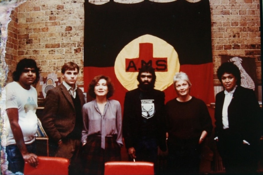 A group of people stand in front of an Indigenous flag with the words AMS. 