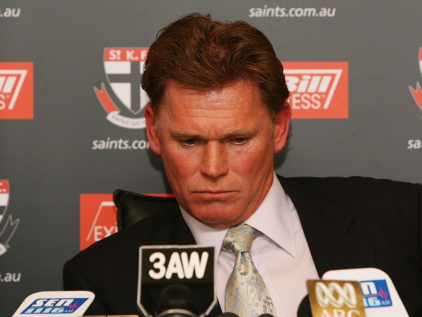 A serious-looking AFL club president looks down as he sits in front of a row of microphones.