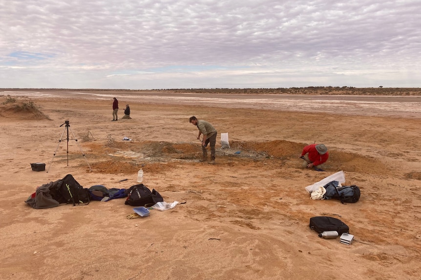 A wide shot of a plain dirt ground with a shallow hole dug by men