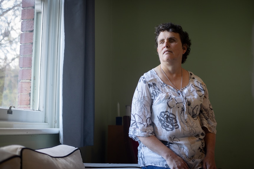 A blind woman sitting on the arm of a couch looking out the window.
