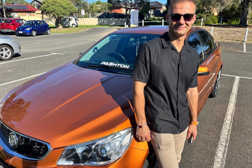 A man wearing a black shirt and chinos standing next to an orange Holden car.