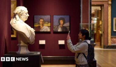 A young woman with black hair, glasses and a backpack stands in front of a statue of a bust and takes a photo. Portraits line the wall behind her.