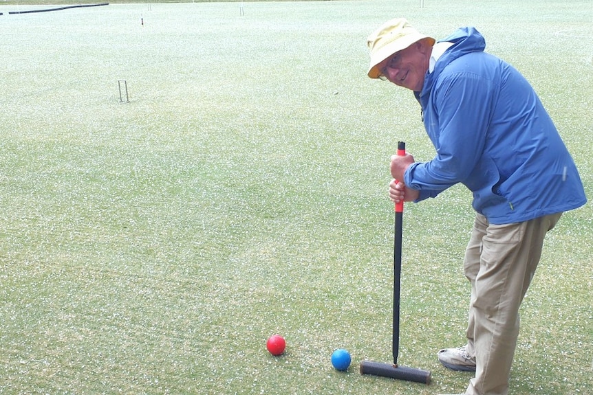 Man about to hit blue ball into red ball using croquet wooden mallet, wearing blue coat and yellow hat smiling at camera