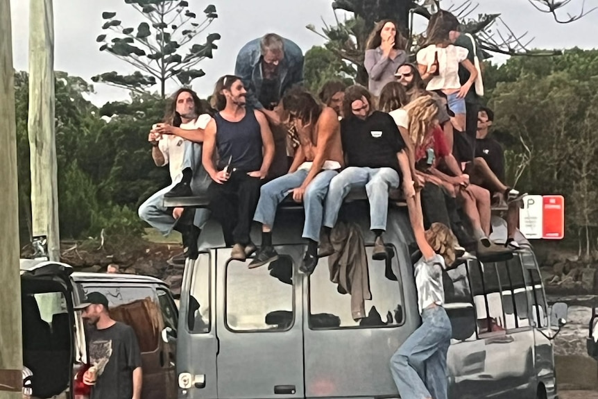 About 12 young men and women drink beers and smoke while sitting on top of a van parked near the ocean.