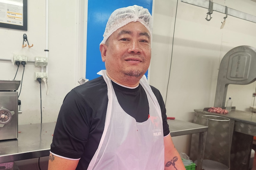 A smiling man wearing an apron and a hair cover, standing at a stainless steel butcher's bench.