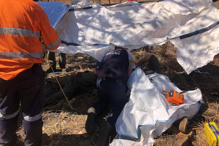 A person in a blue shirt kneels over a person lying on the ground under a white tarp