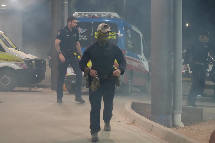 A police officer wearing a bandana over his mouth and nose with a baseball cap on at a hospital ambulance ramp.