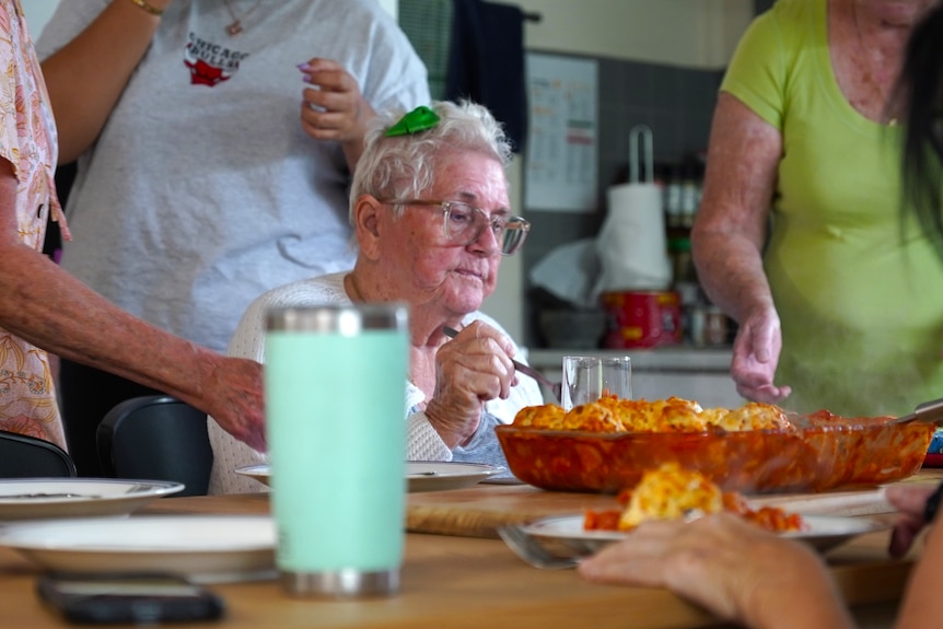 An elderly lady eats her lunch at a dining table. She is surrounded by other women.