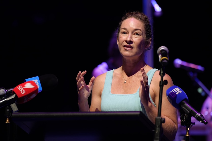 A woman in a light blue top with her hair up talks into media microphones inside a theatre.