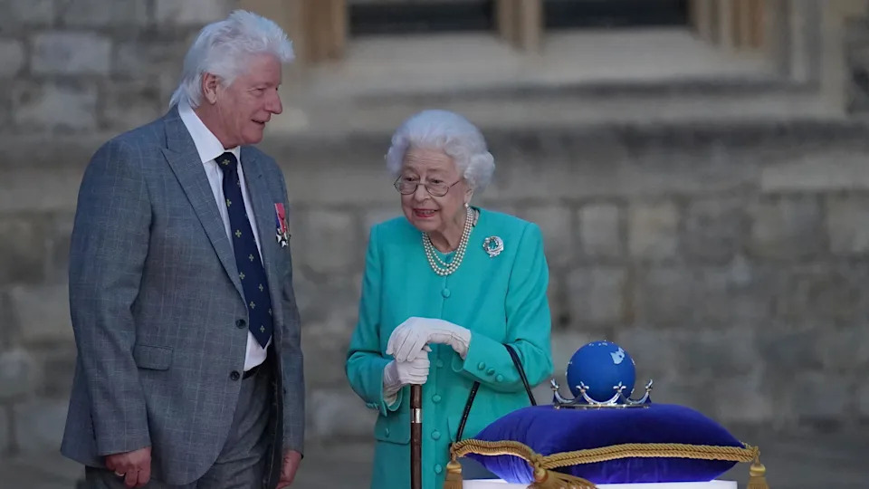 Bruno Peek standing next to Queen Elizabeth II outside. Peek is wearing a grey suit and is looking towards Queen Elizabeth II. She is dressed in a teal blue outfit with pearl necklaces and she is wearing white gloves. They are standing in front of a purple cushion which has the Platinum Jubilee 'Commonweath of Nations Globe' on it.