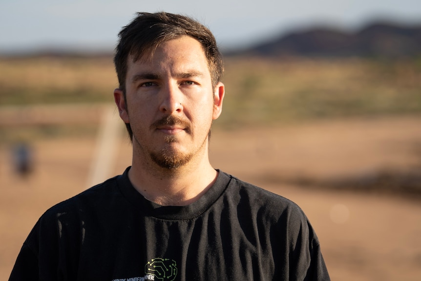 A young man with brown hair, beard, and goatee looks into camera with a neutral expression.