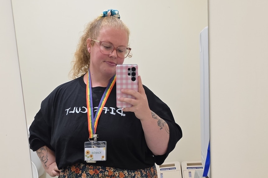 a red haired woman in a black shirt and rainbow lanyard takes a selfie in a mirror