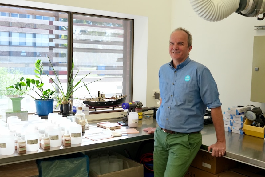 A man smiles as he leans on a bench inside a laboratory.