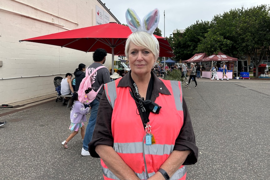 A woman wears a hi-vi vest and Easter bunny ears in front of an outdoor market with pop-up marquees next to a large building