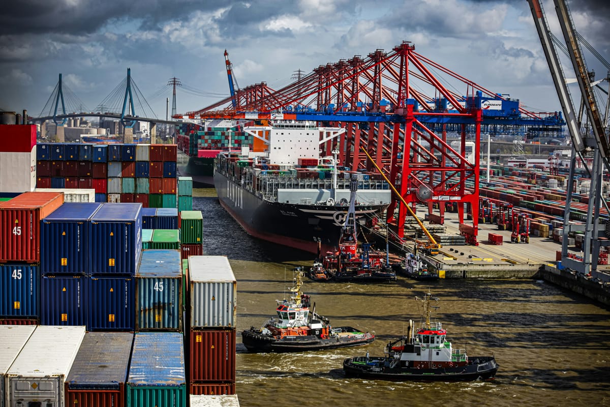 Vibrant port scene with a large cargo ship being guided by tugboats, flanked by towering red cranes and stacks of multicolored shipping containers under a dynamic sky (Bernd Dittrich/Unsplash)