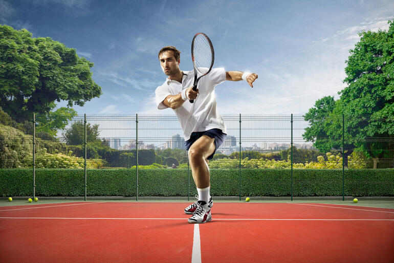 Male tennis player in action, hitting a forehand on an outdoor court, dynamic sports shot with racket motion, fenced court surrounded by greenery and city skyline in the background.