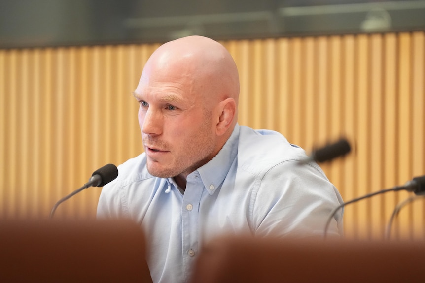 A middle-aged bald man in a pale shirt leans into a microphone.