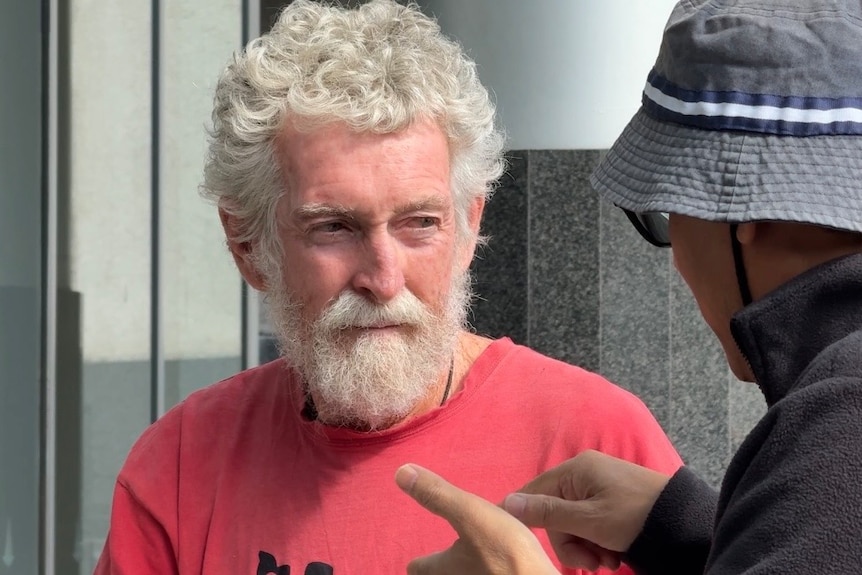 A man with grey hair and beard in a red tshirt talking to a person with their back to the camera