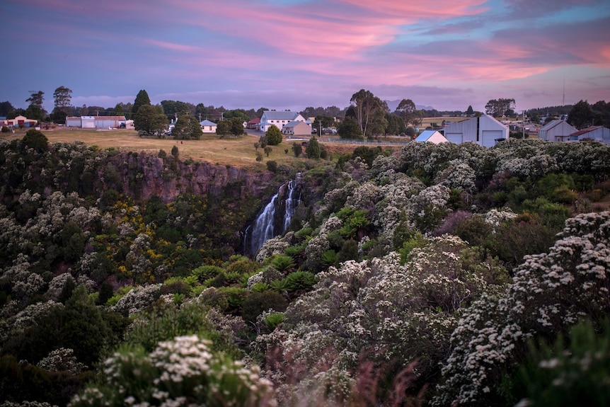 Purple, blue and pink skies over a tree-lined gorge. Houses are in the background and a waterfall can be seen