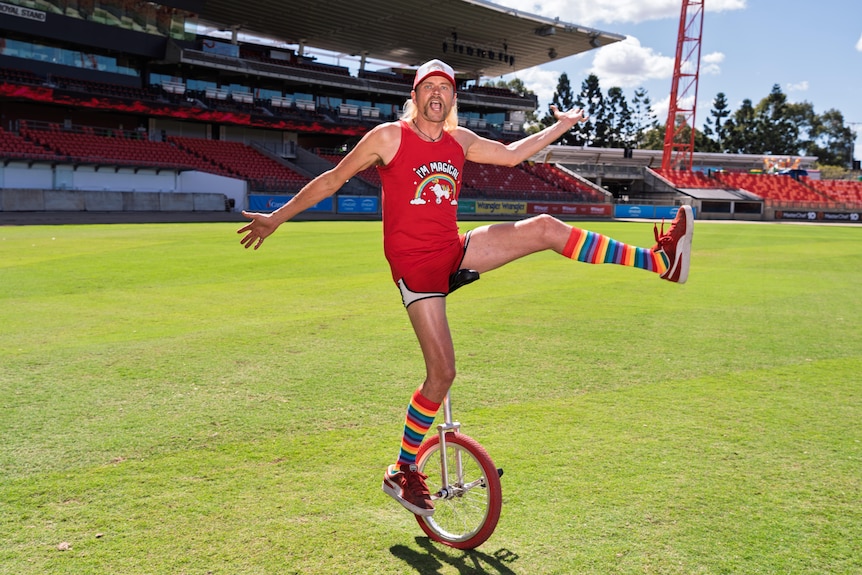 Jamey pulls a funny face and waves his hand and one leg while riding a unicycle.