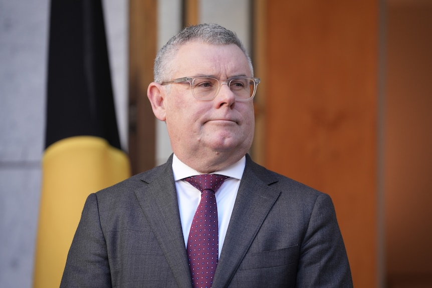 A man in suit, tie and glasses standing in front of a furled flag.