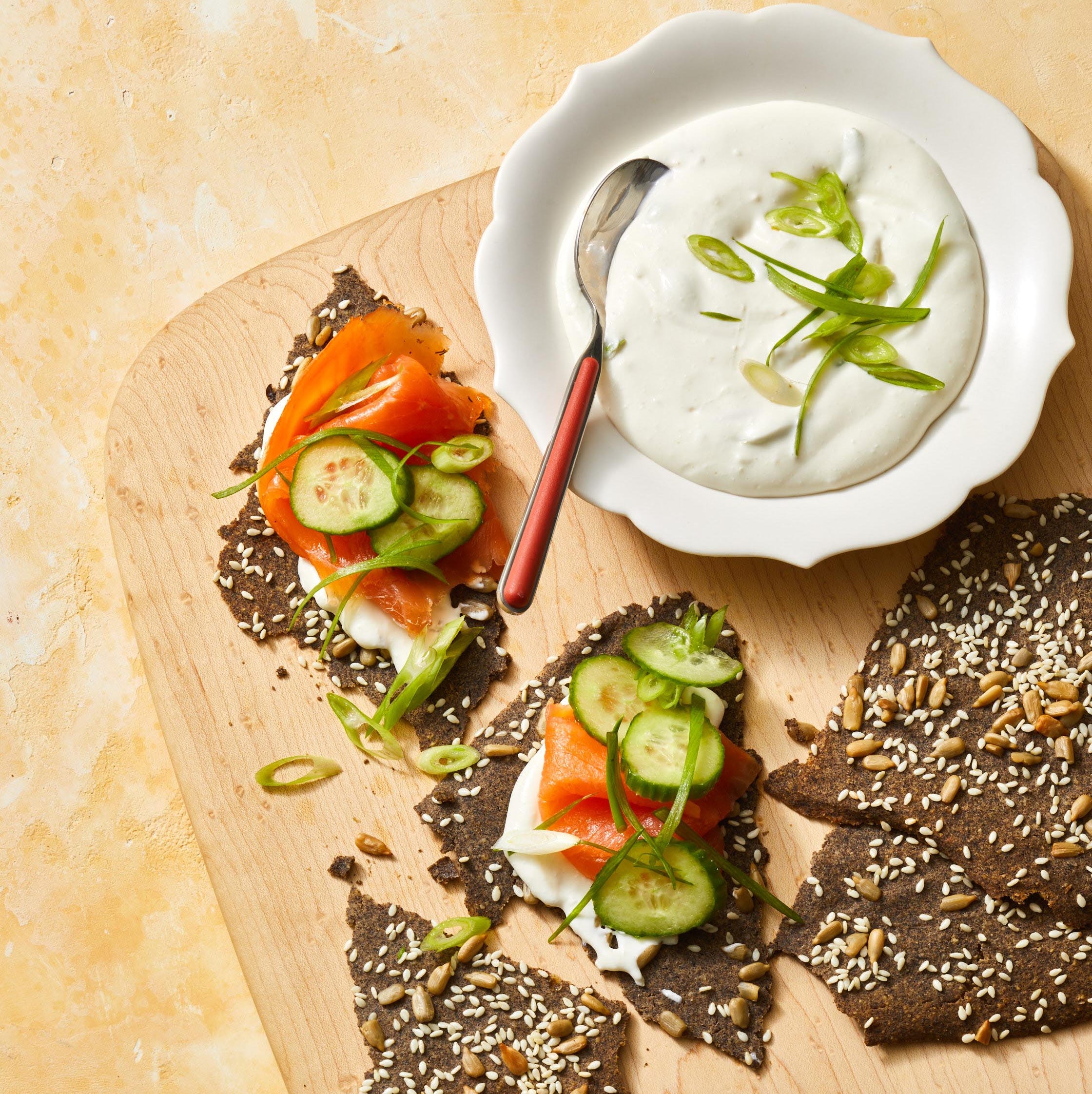 a wood food board with buckwheat crackers and smoked salmon and cucumbers on top