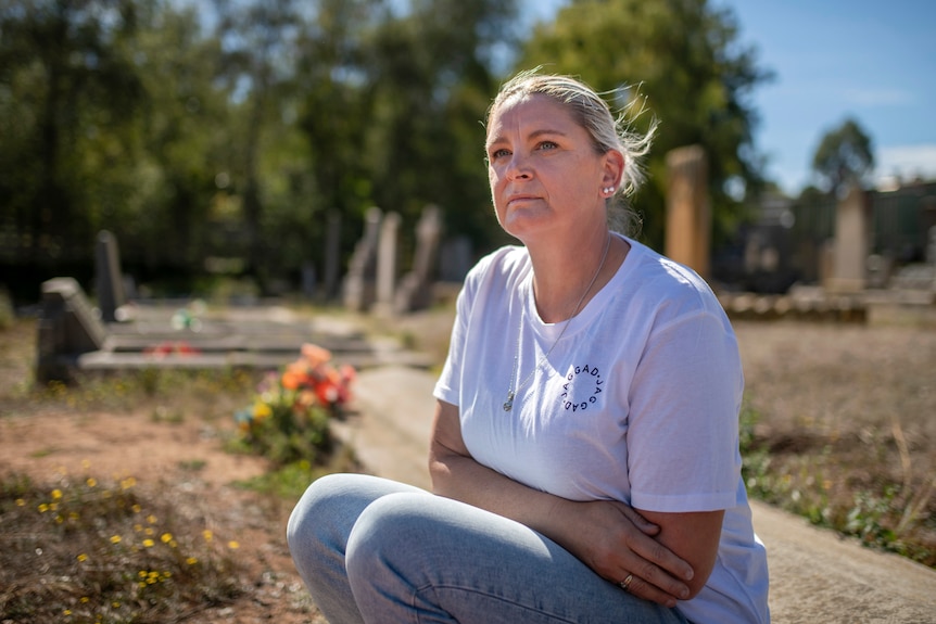 A woman in a white t-shirt and blue jeans sits by a grave in a cemetery with green trees and flowers in the backgound.
