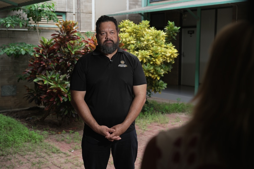 A man in black polo shirt stands in a courtyard and speaks to a reporter.