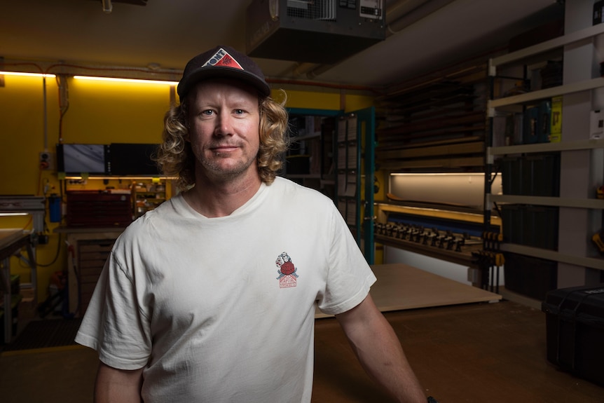 A man in a shirt and hat, standing in a workshop.