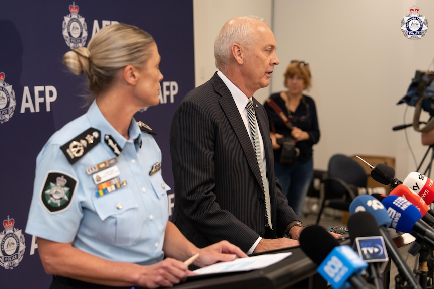 Ross wears a suit jacket and speaks into a row of microphones.