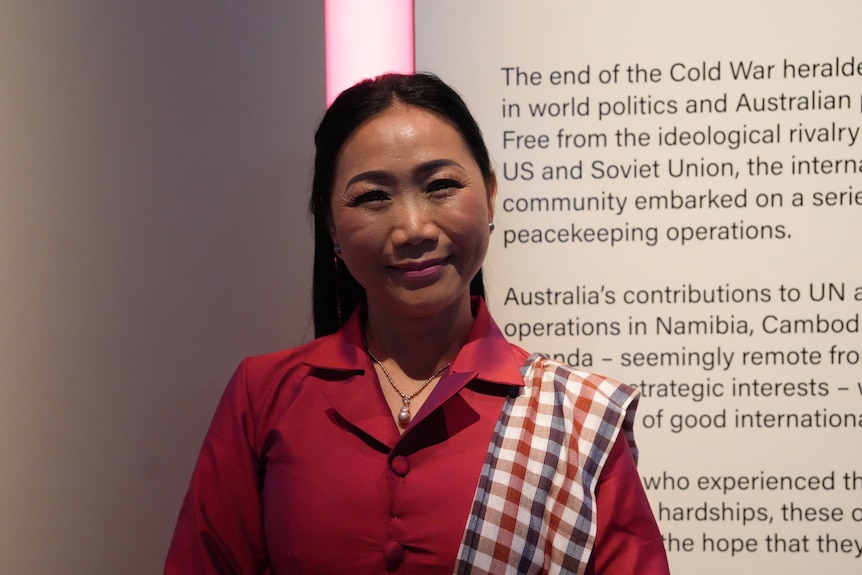 A woman with long dark hair wearing a red shirt stands in a museum smiling.