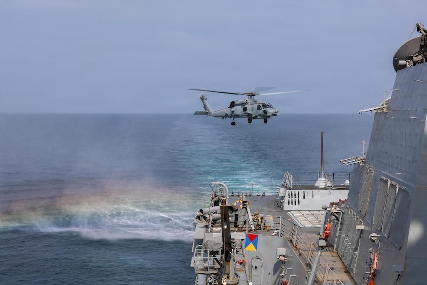 A helicopter launches from the deck of a military ship.