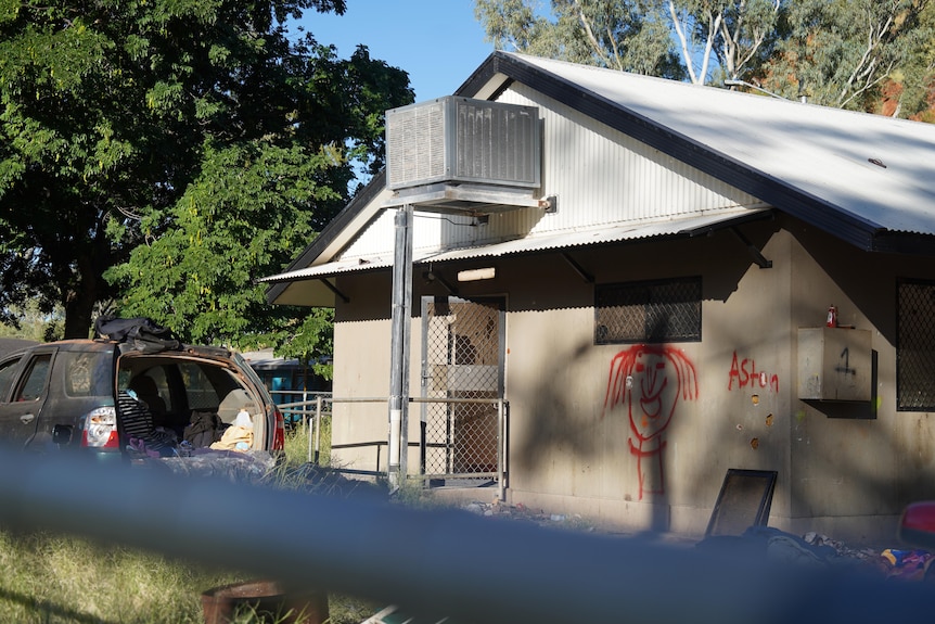 A concrete house with an open car in front, graffiti on the walls and rubbish on the ground