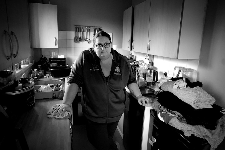 A woman standing in a narrow kitchen with a pile of laundry to her left and a sink to her right.