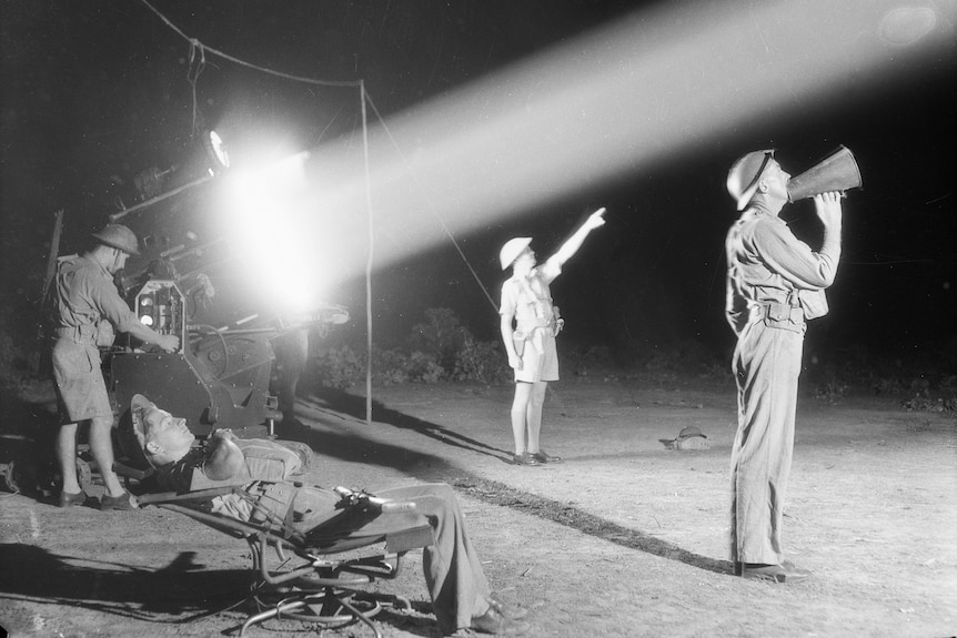 An army crew operating a large searchlight, the uniformed men are looking towards and pointing up at the night sky.