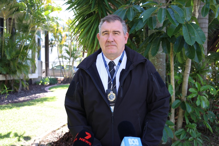 A man with a police badge around his neck a shirt and a black jacket with trees behind him.