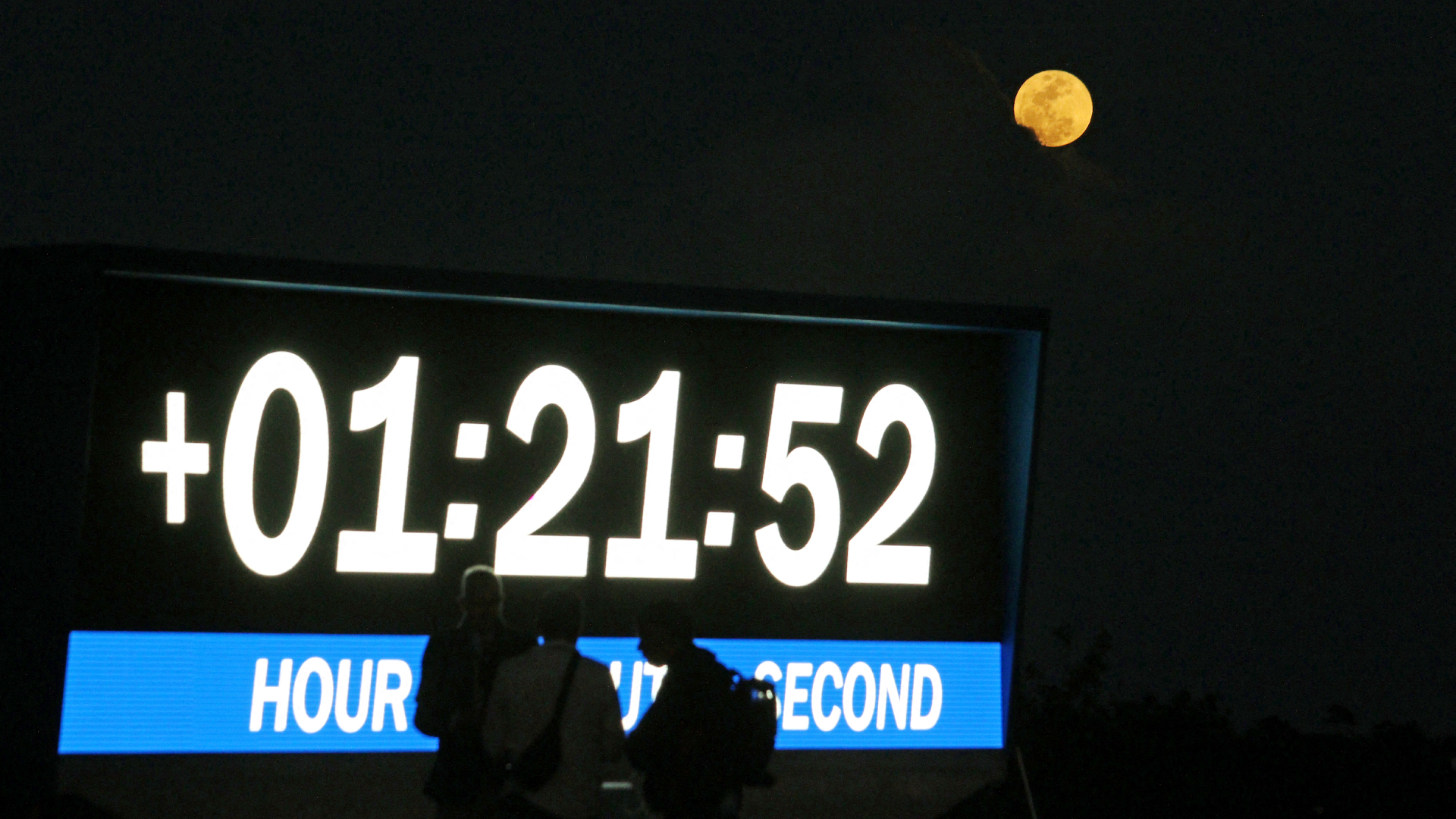 A full moon shines in a night sky above a launch countdown board at the Kennedy Space Center, with people standing in the foreground.