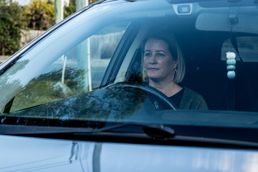 A middle-aged woman with shoulder length blonde hair sitting at the front wheel of a car.