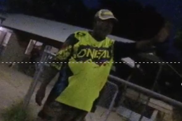 A low-res dark image, of a man in a bright yellow shirt and cap, raising his hand in the air, with a house in background