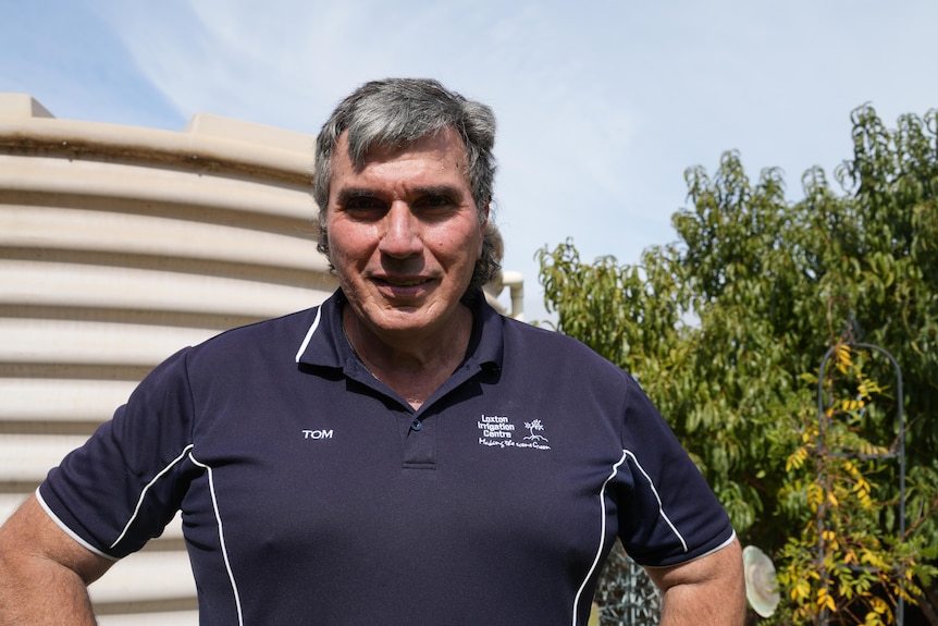 A grey-haired man in a branded polo shirt stands near a water tank and some trees.